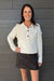 A woman shown wearing a cream cable-knit sweater and a dark brown mini skirt, standing against a gray wood-paneled wall.