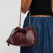 A dark burgundy woven leather shoulder bag with a gold chain strap, shown being worn by a model in a black top and blue skirt against a light gray background.