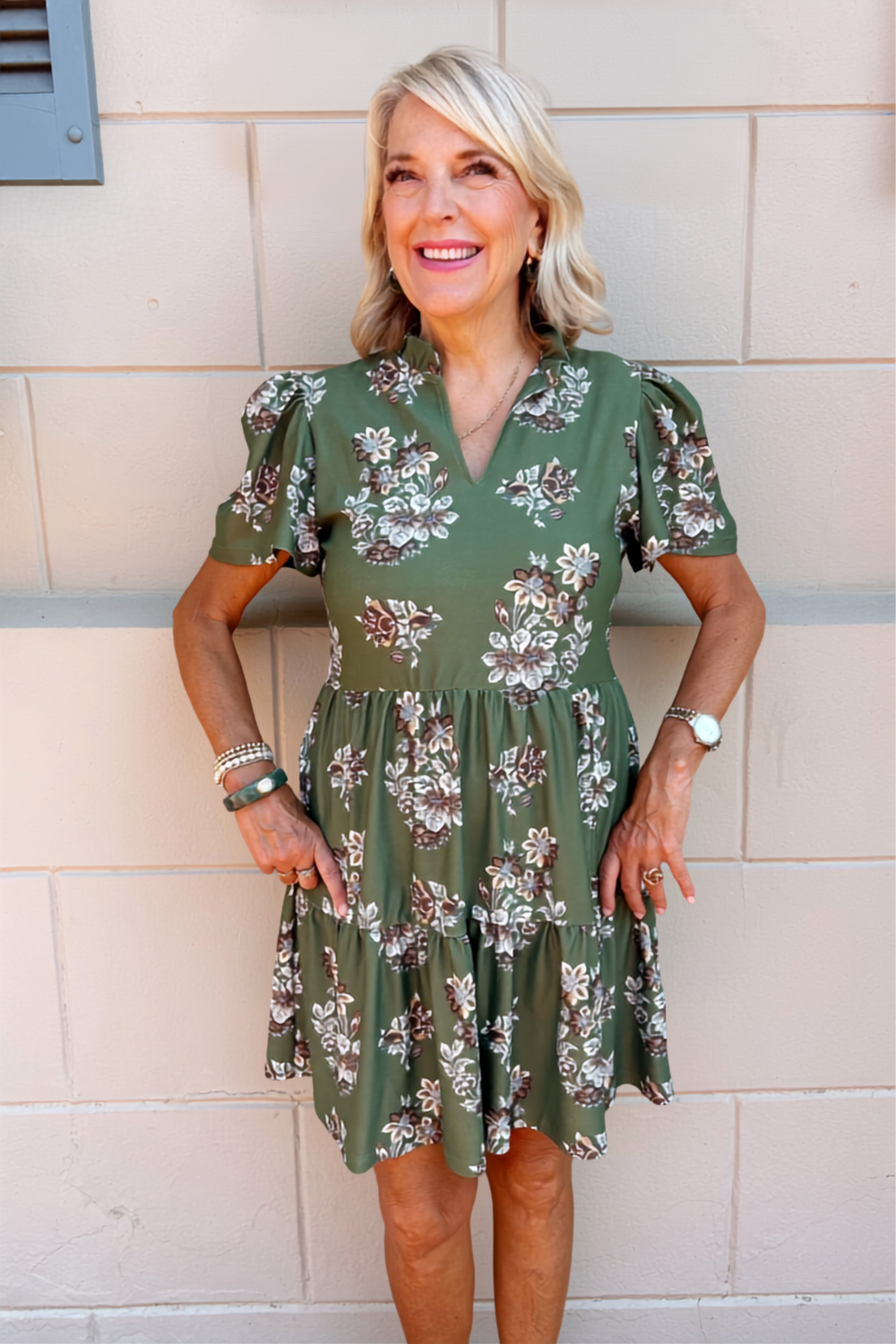 A smiling blonde woman wearing an olive green short-sleeve dress with a white and brown floral pattern, standing against a beige brick wall.