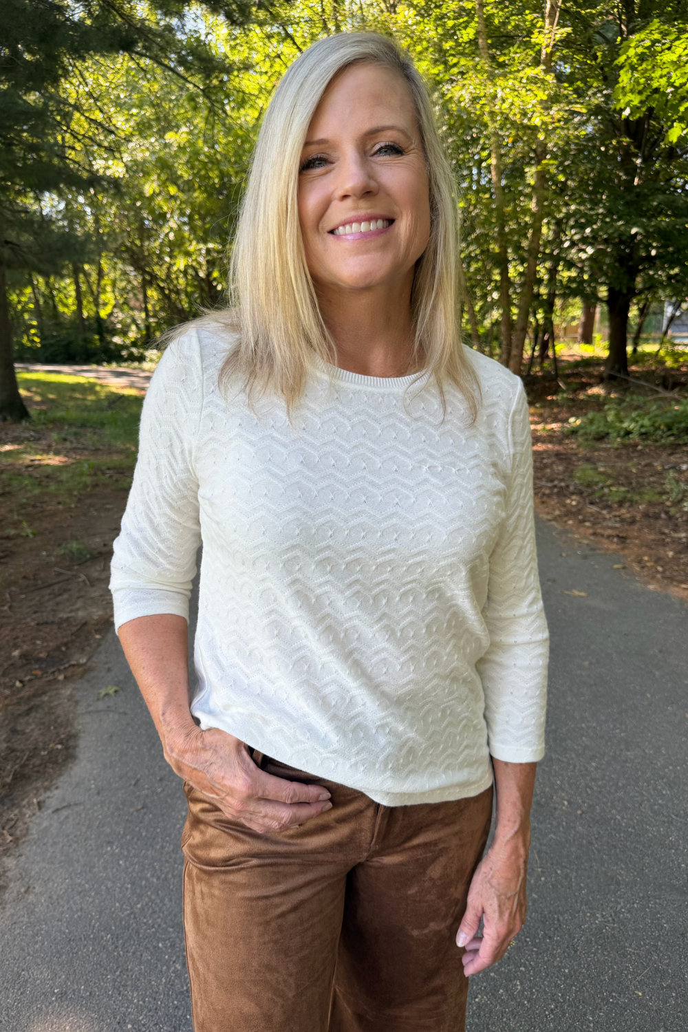 A smiling blonde woman wearing a cream-colored textured knit sweater and brown pants, standing on an outdoor paved path surrounded by green trees.