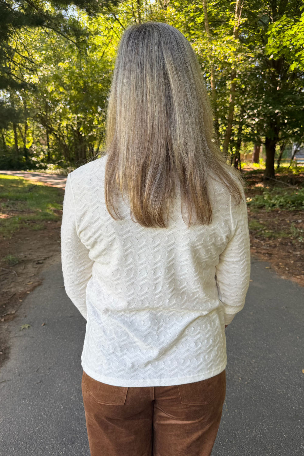 A woman viewed from behind wearing a cream-colored textured knit sweater and brown pants, standing on an outdoor paved path surrounded by green trees.