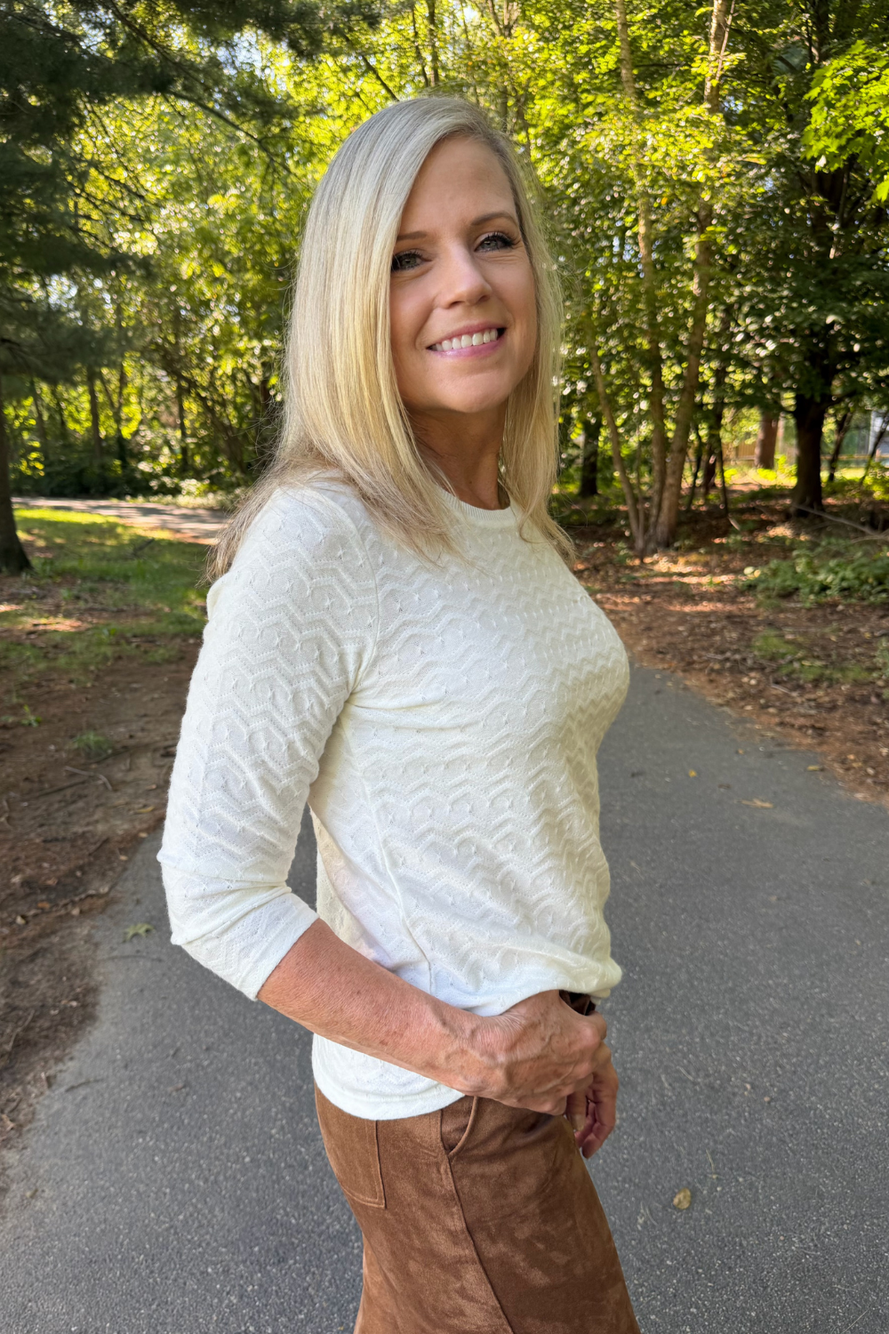 A smiling blonde woman wearing a textured cream-colored long-sleeve top and brown pants, standing on an outdoor paved path surrounded by green trees.