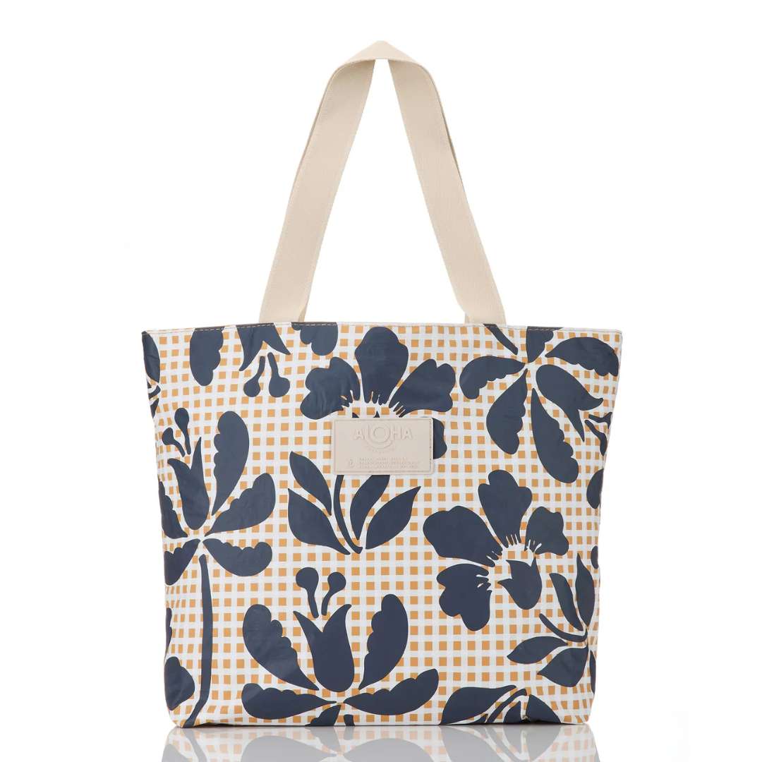 A canvas tote bag featuring a navy blue floral print over a warm beige and orange gingham check pattern, with cream-colored shoulder straps and a small leather brand patch on the front.