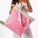 A woman holds a large tote bag with a pink and white geometric pattern and beige handles against a clean white background.
