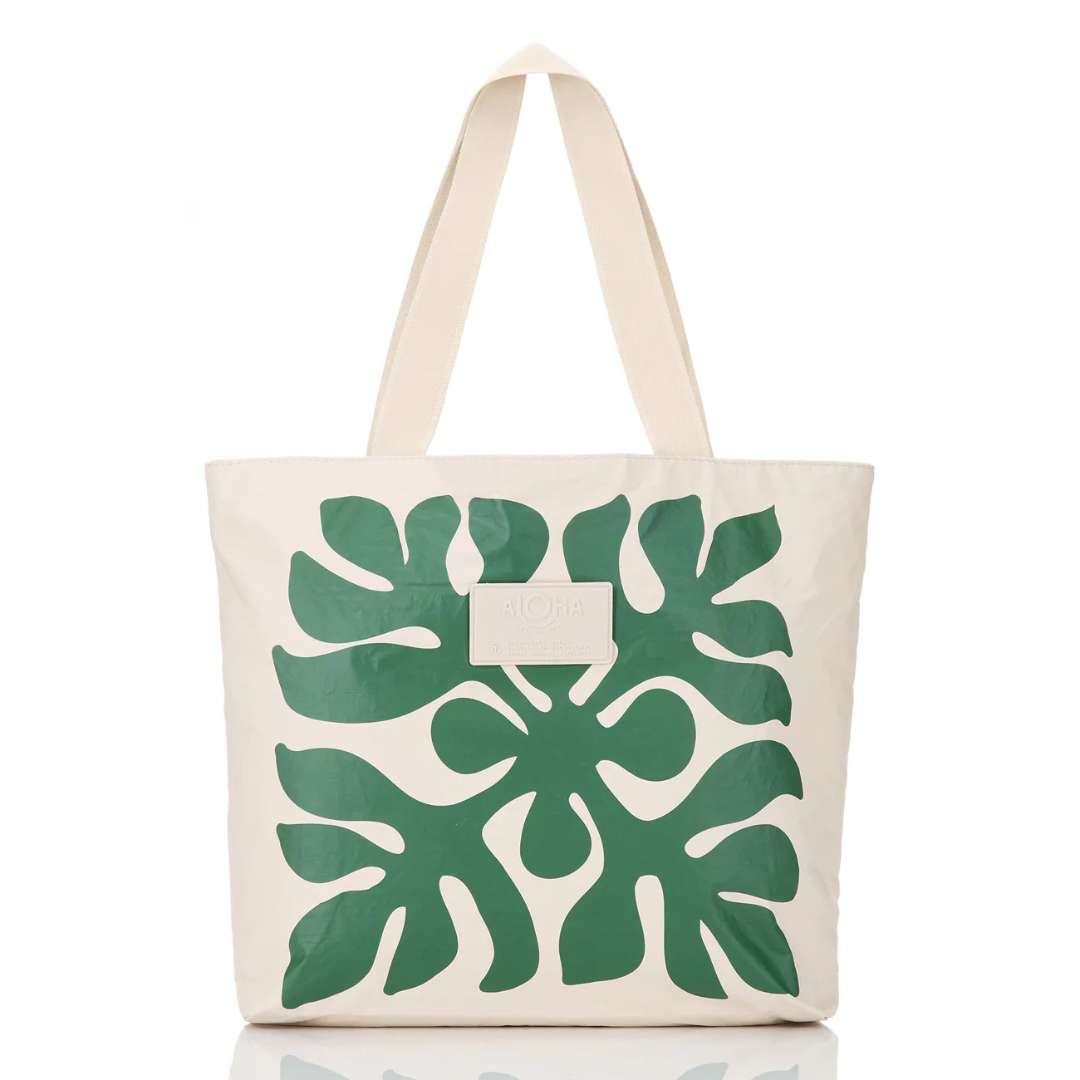 An off-white canvas tote bag with a large green monstera leaf pattern printed on the front, featuring dual shoulder straps and a small white brand patch.