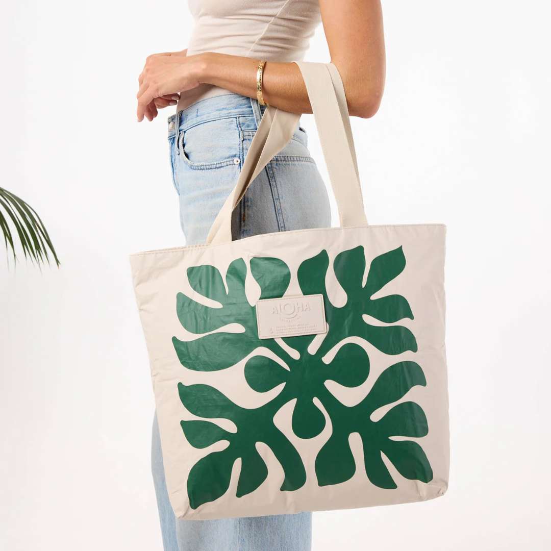 A person wearing a white top and light blue jeans holds a cream canvas tote bag with a large green monstera leaf pattern. The bag has a small white brand label that reads 'MOON'.