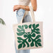 A person wearing a white top and light blue jeans holds a cream canvas tote bag with a large green monstera leaf pattern. The bag has a small white brand label that reads 'MOON'.