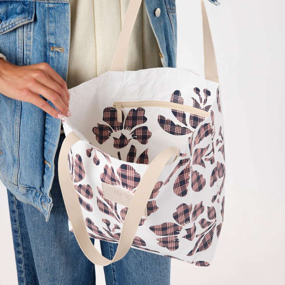 A person holding a white tote bag with a floral plaid pattern and beige shoulder straps. The person is wearing a denim jacket and jeans against a plain white background.
