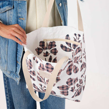 A person holding a white tote bag with a floral plaid pattern and beige shoulder straps. The person is wearing a denim jacket and jeans against a plain white background.