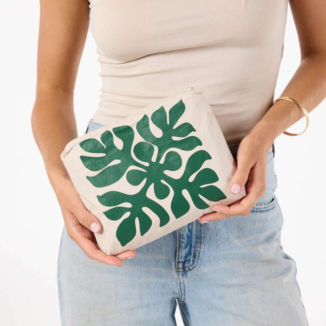 A person in a beige ribbed tank top and light blue jeans holds a canvas clutch bag with a large green monstera leaf pattern on a white background.