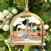 An arched Christmas ornament featuring a colorful miniature illustration of an Annapolis harbor scene with a sailboat, buildings, and a gold-toned border, hanging among green tree branches and white glitter ornaments.