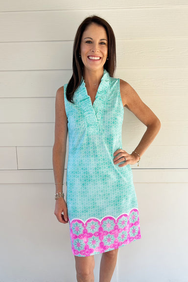 A woman modeling a sleeveless mint green and white patterned tunic dress with a pink and white floral scalloped hem, standing against a white paneled wall.