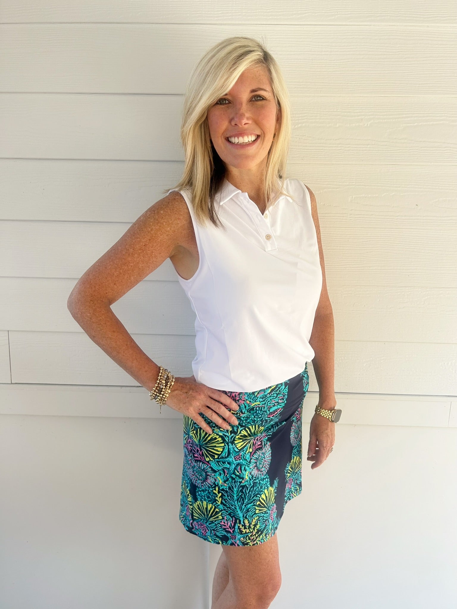 A smiling blonde woman stands against a white paneled wall wearing a white sleeveless collared top and a colorful tropical print mini skirt with teal, green, yellow, and purple tones.