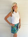 A smiling blonde woman stands against a white paneled wall wearing a white sleeveless collared top and a colorful tropical print mini skirt with teal, green, yellow, and purple tones.
