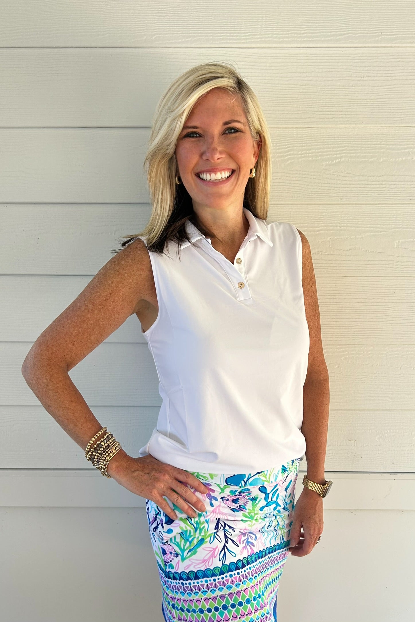 A smiling blonde woman stands against a white paneled wall, wearing a sleeveless white polo shirt and a colorful patterned skirt with blue, green, pink, and white botanical and geometric designs.