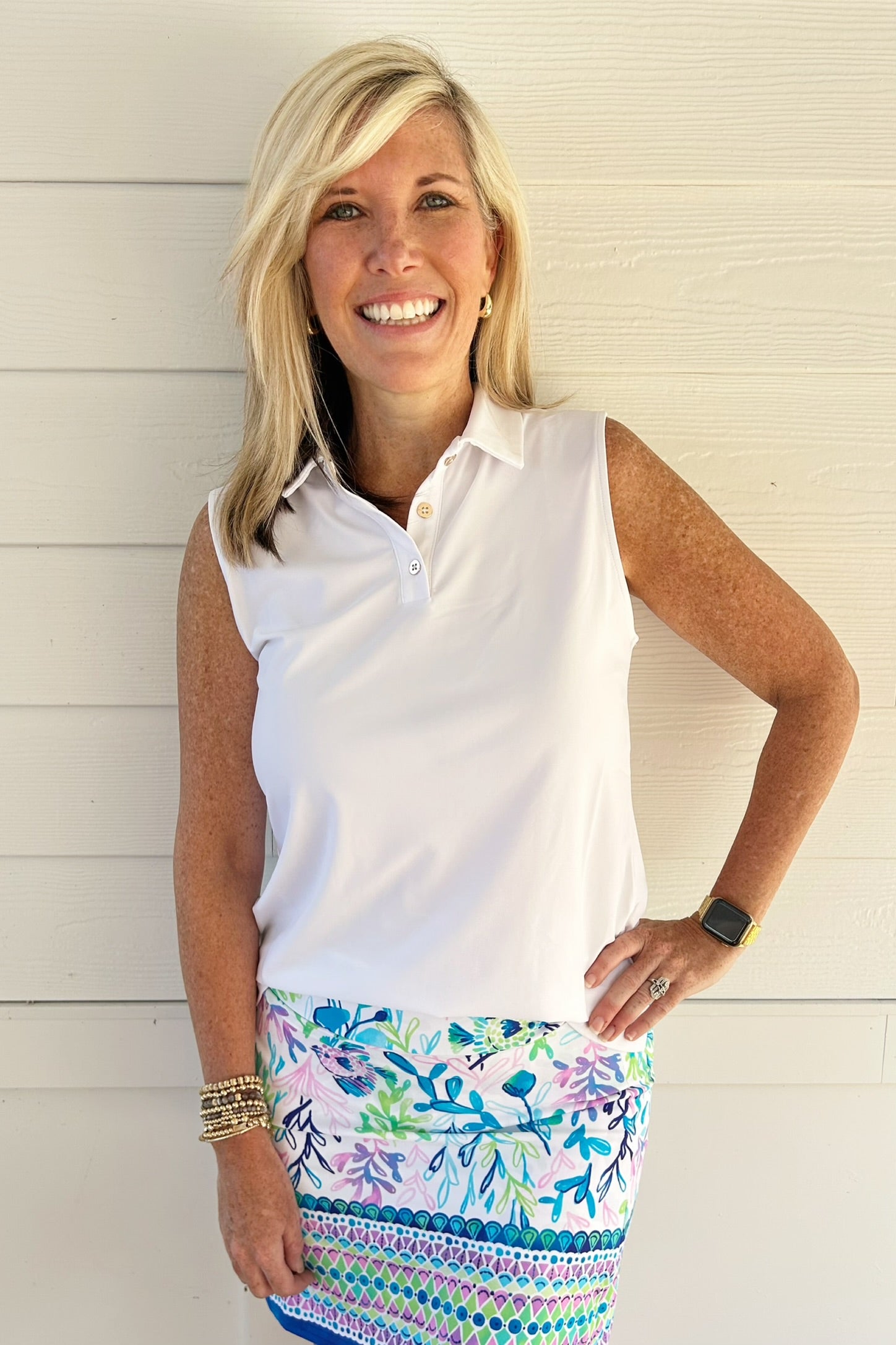 A smiling blonde woman wearing a white sleeveless polo shirt and a colorful patterned skirt, standing against a white wood-paneled wall.