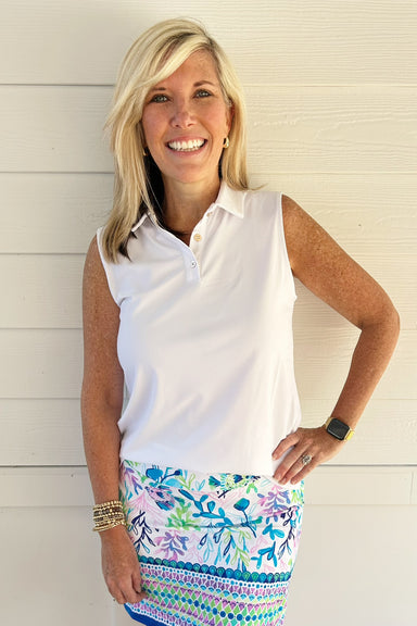 A smiling blonde woman wearing a white sleeveless polo shirt and a colorful patterned skirt, standing against a white wood-paneled wall.