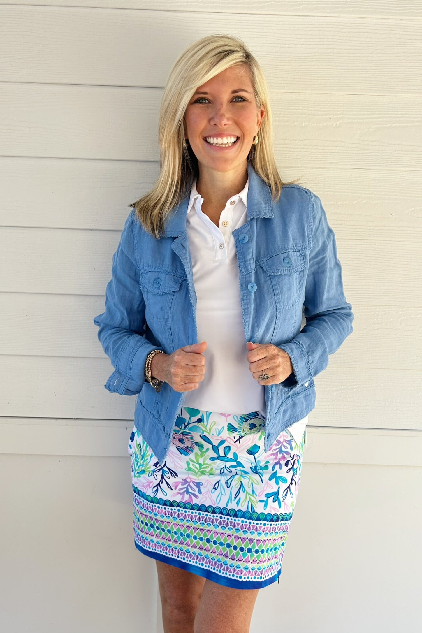 A smiling blonde woman stands against a white paneled wall wearing a light blue denim jacket over a white collared shirt and a white mini skirt with a colorful blue, green, and purple floral and geometric pattern.