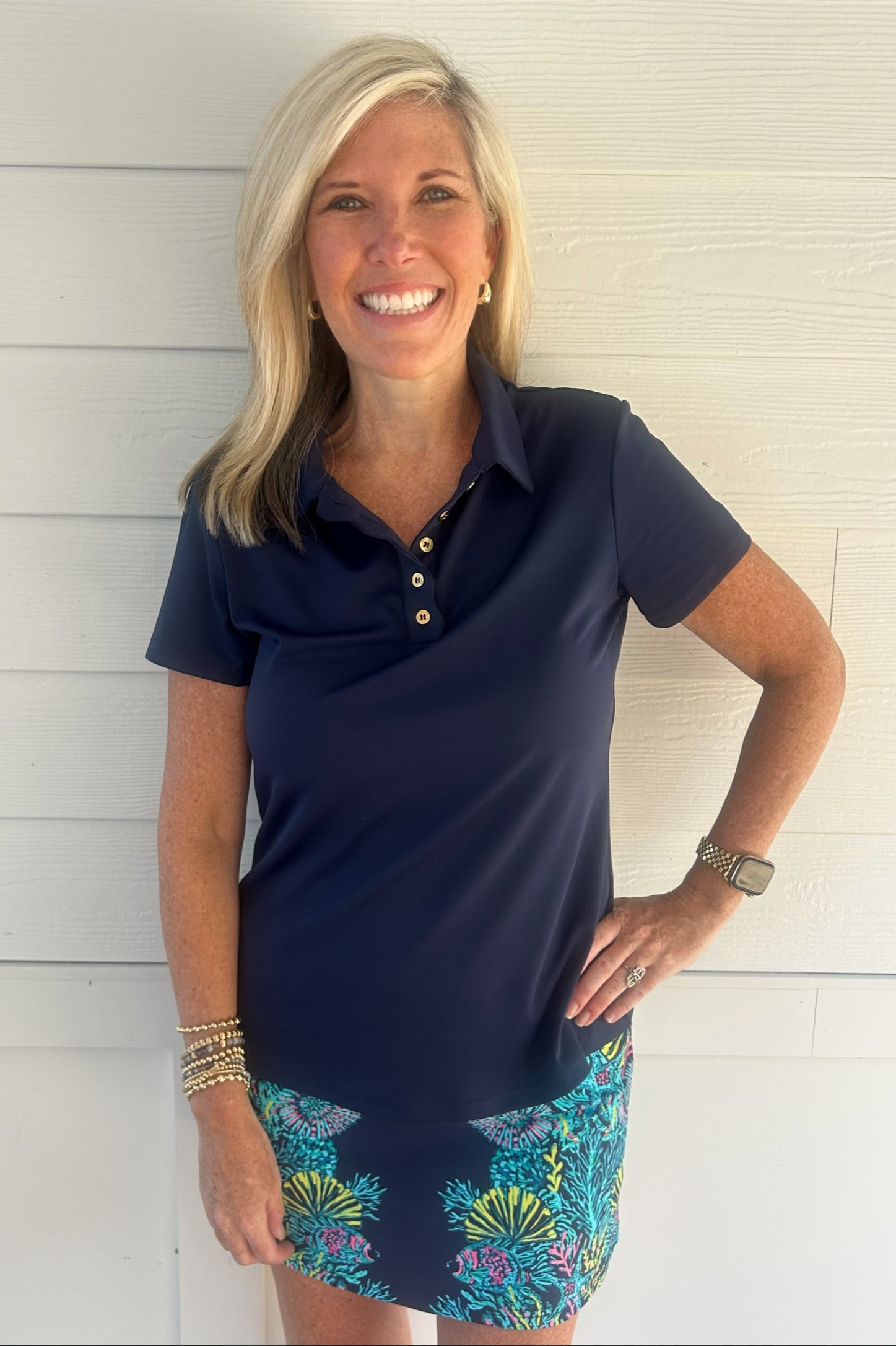 A smiling blonde woman wearing a navy blue short-sleeve polo shirt with gold buttons and a colorful tropical print skirt, standing against a white shiplap wall.