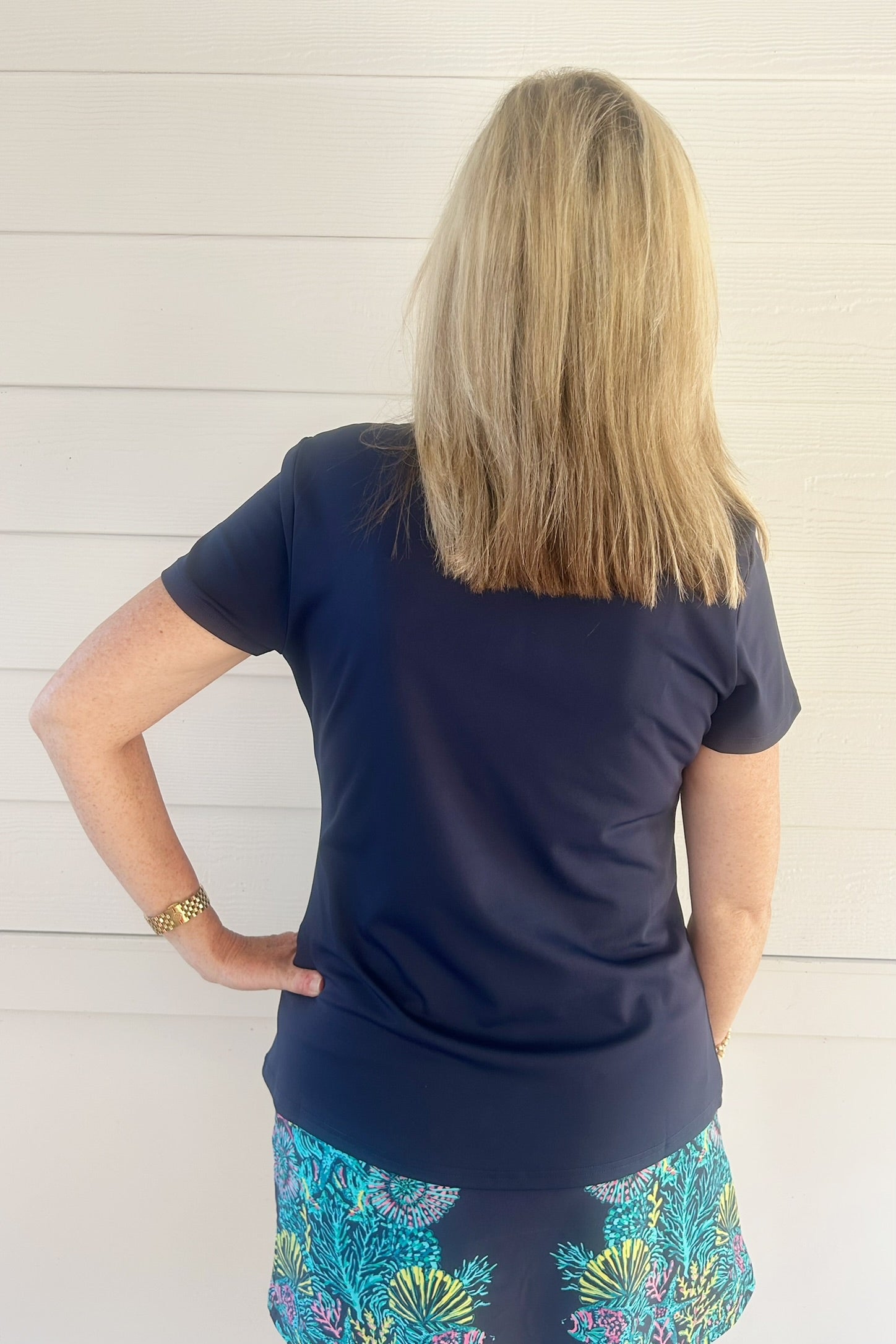 A woman viewed from behind wearing a navy blue short-sleeve top and a colorful blue patterned skirt, standing against a white paneled wall.