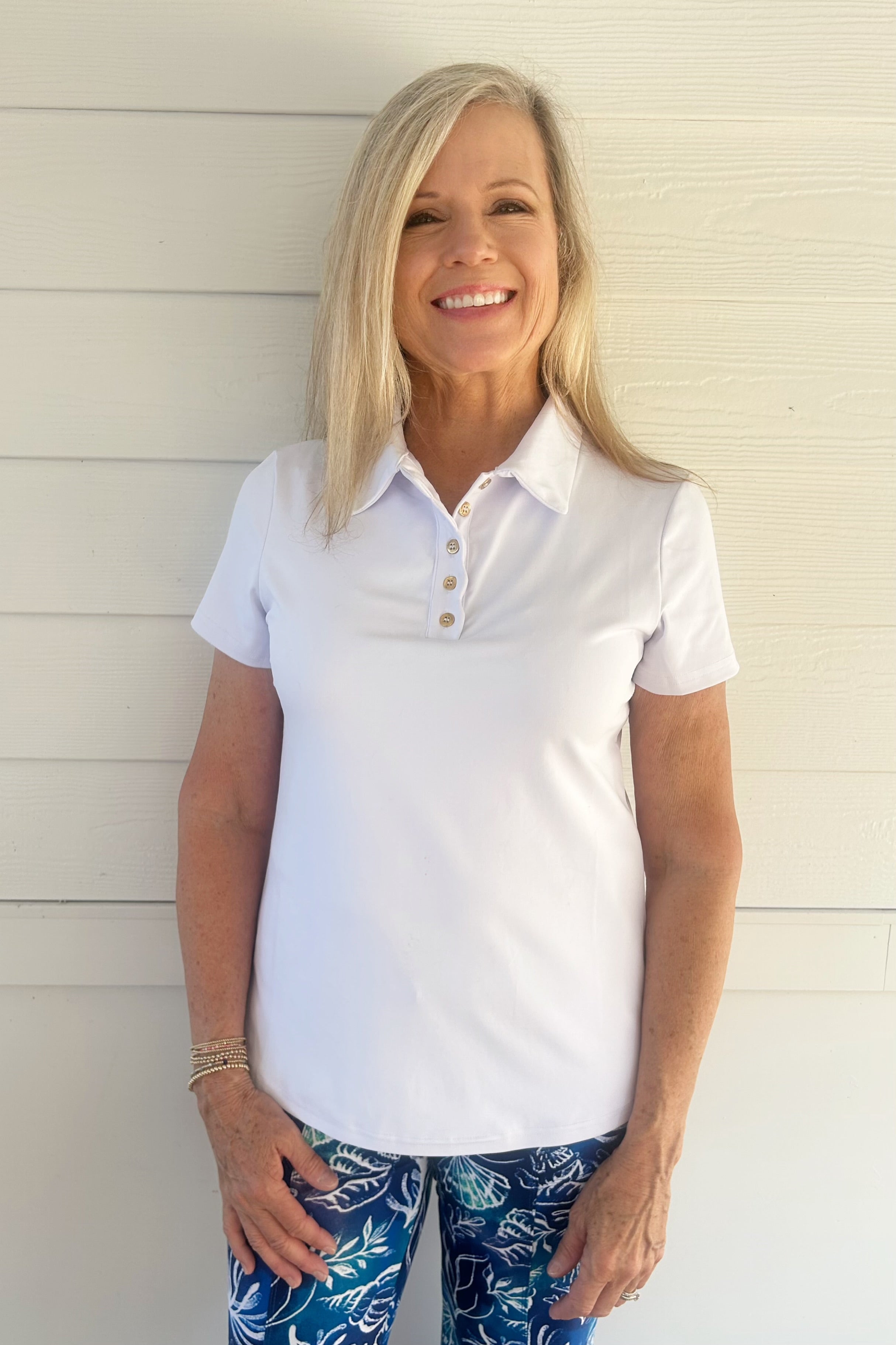 A smiling blonde woman wearing a white short-sleeve polo shirt and blue patterned leggings, posing against a white wood-paneled wall.