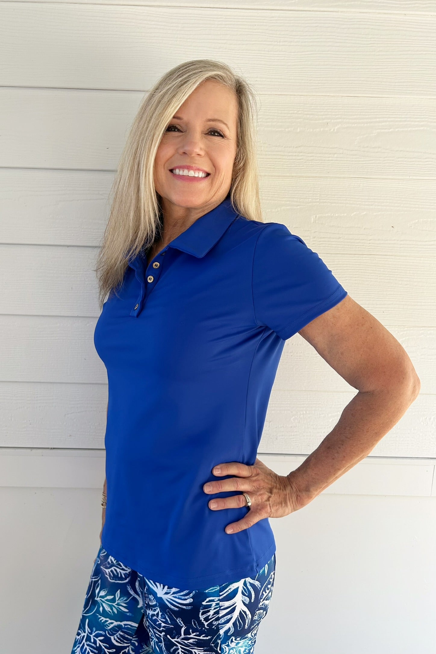 A smiling blonde woman wearing a bright royal blue short-sleeve polo shirt and blue patterned bottoms, standing against a white shiplap wall.
