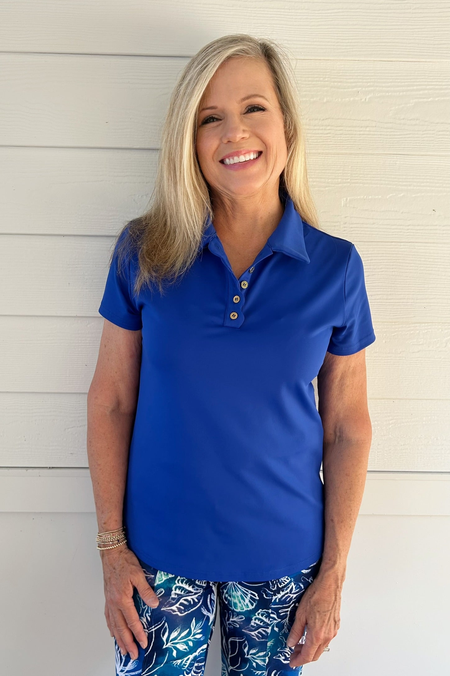 A smiling blonde woman wearing a bright royal blue short-sleeve polo shirt with a three-button placket and matching blue patterned pants, standing against a white horizontal siding wall.