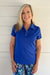 A smiling blonde woman wearing a bright royal blue short-sleeve polo shirt with a three-button placket and matching blue patterned pants, standing against a white horizontal siding wall.