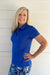 A smiling blonde woman wearing a bright royal blue short-sleeve polo shirt and blue patterned bottoms, standing against a white shiplap wall.