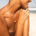 A close-up lifestyle shot of a woman on a sandy beach wearing a delicate gold chain necklace with a small white bead pendant.