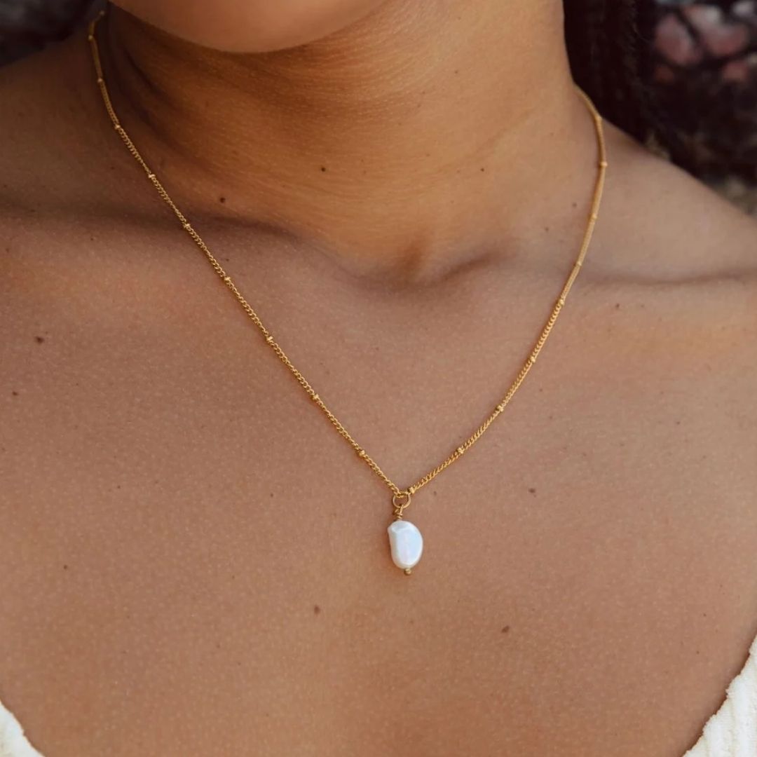A close-up of a model's neck and chest wearing a delicate gold chain necklace with a small, irregular white pearl pendant.