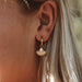 A close-up of a person's ear wearing a gold hoop earring with a dangling fan-shaped charm and a small pearl-like center, against light hair and a softly blurred background.