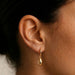 A close-up of a person's ear wearing a small gold hoop earring with a dangling teardrop-shaped gold charm.