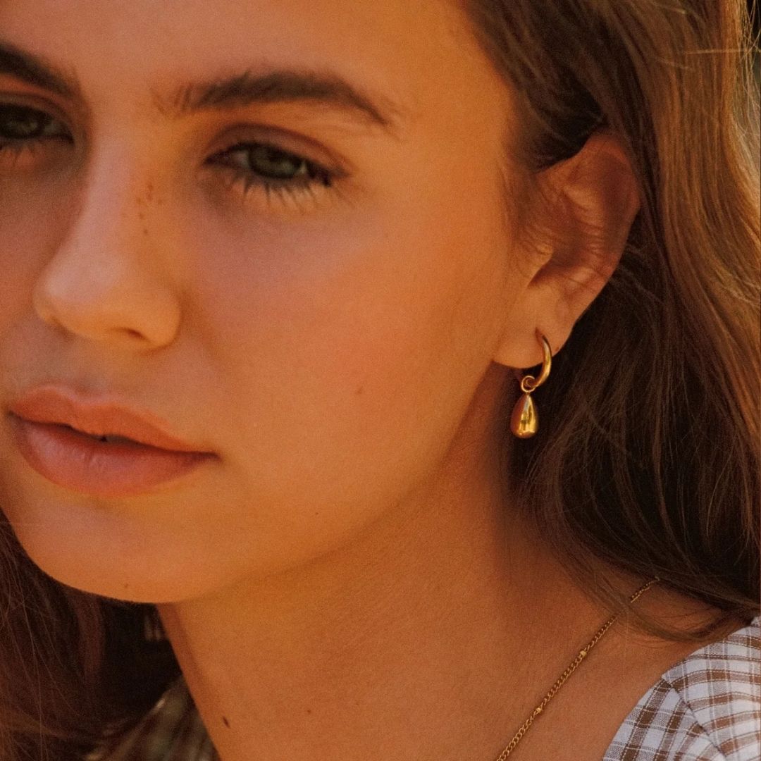 A close-up of a woman wearing a small gold hoop earring with a teardrop-shaped drop, shown in warm natural light.