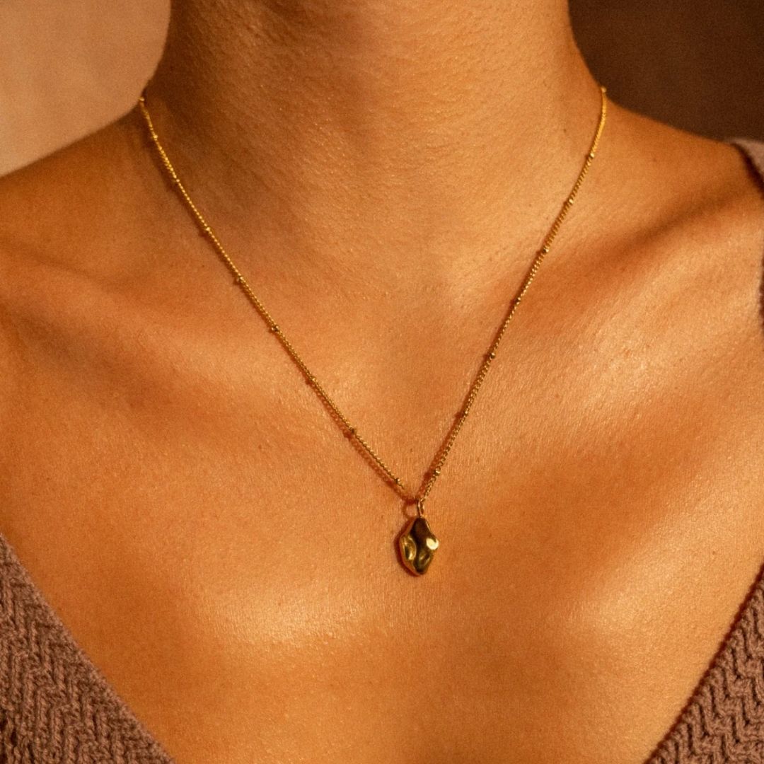 A gold chain necklace with a small teardrop-shaped gold pendant worn on a model's neck, shown in warm, golden lighting.