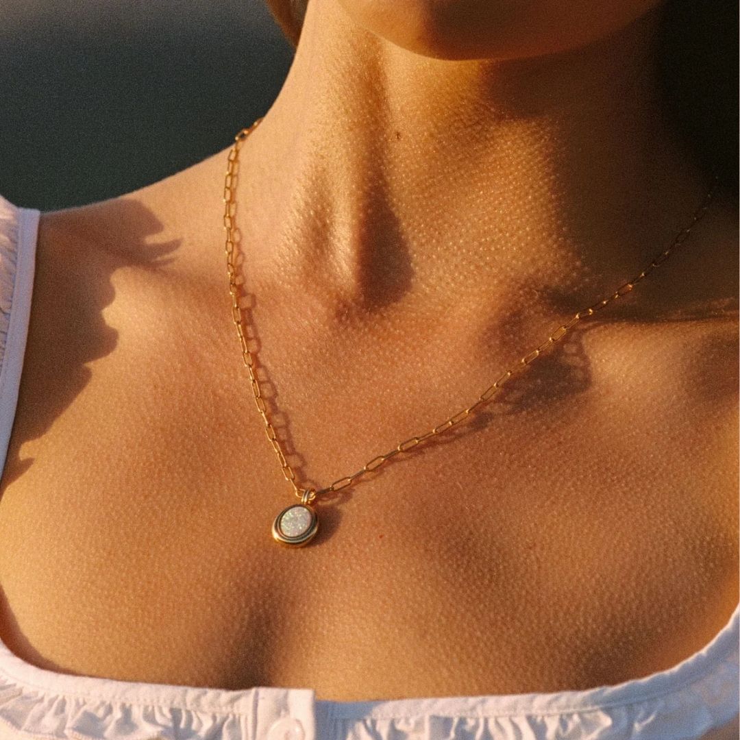 A close-up lifestyle shot of a woman's neck and chest wearing a delicate gold chain necklace with a small oval pendant featuring a white center and gold border.