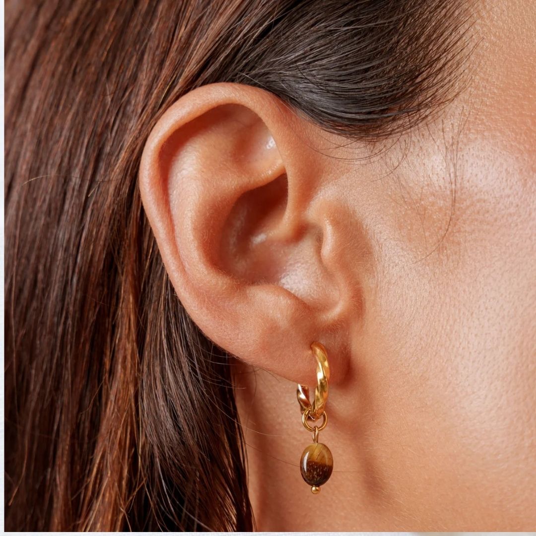 A close-up of a person's ear wearing a small gold hoop earring with a dangling polished brown stone.