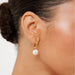 A close-up of a model's ear wearing a delicate gold huggie hoop earring with a dangling textured white flower charm.
