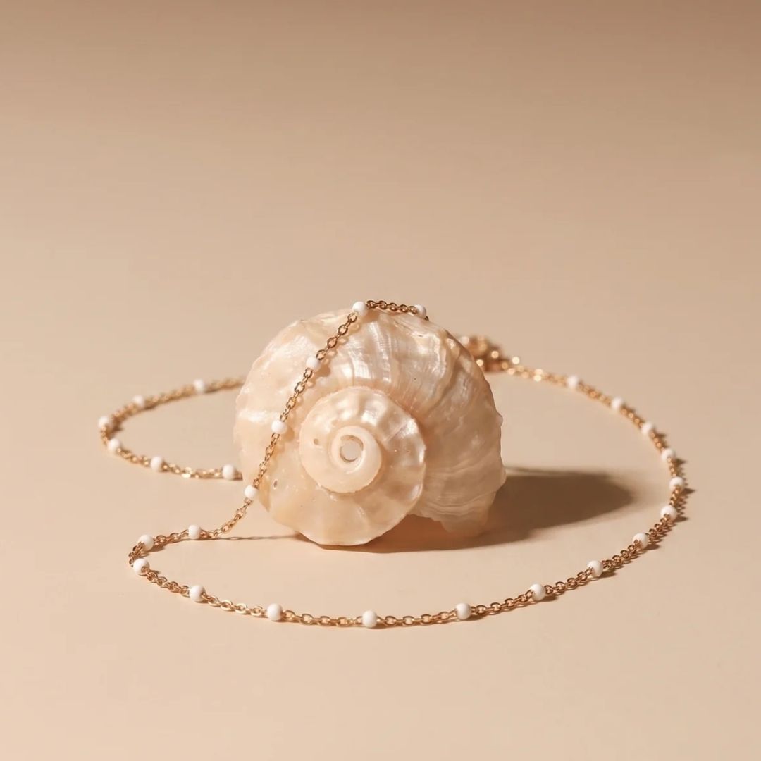 A shell-shaped crossbody bag with a cream-colored mother-of-pearl shell body and a delicate gold chain strap, displayed on a warm beige background.