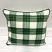 A square throw pillow with a green and off-white plaid pattern and black piping, resting on a white wooden floor against a light gray wall.
