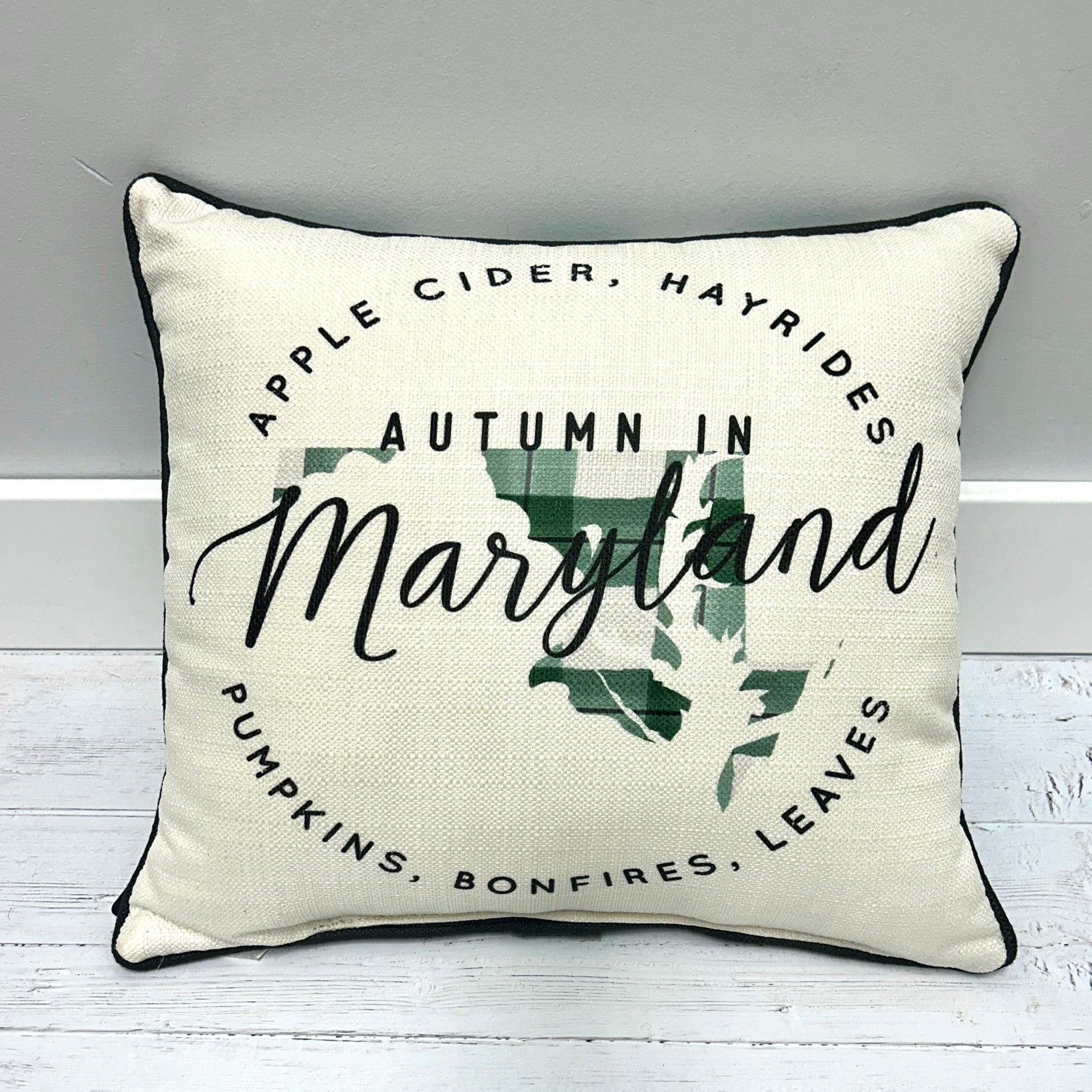 An off-white textured throw pillow with black piping, featuring a graphic design that reads 'Autumn in Maryland' with a green plaid map of Maryland and seasonal words like apple cider, hayrides, pumpkins, bonfires, and leaves.