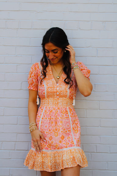 Woman wearing a pink and orange patterned dress against a white brick wall.