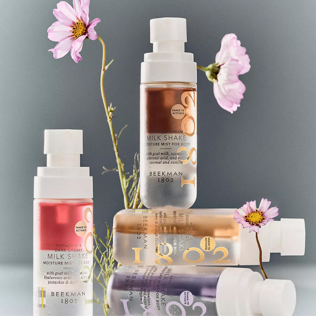 A styled product lineup of BEEKMAN 1803 Milk Shake Dewy Mist for Body and Milk Shake Moisture Lotion, featuring white pump bottles and a squeeze tube in pink, peach, and lavender shades, arranged with pink flowers on a gray background.