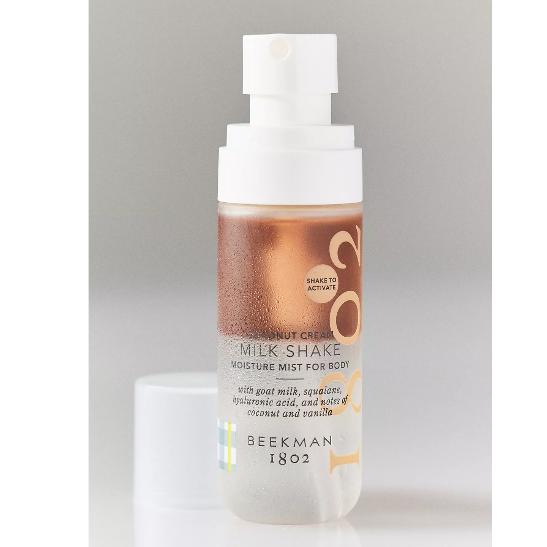 A bottle of Beekman 1802 Coconut Cream Milk Shake Moisturizing Mist for Body, featuring a white spray cap and a frosted bottle showing a coppery rose liquid inside, set against a neutral gray background.