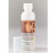 A bottle of Beekman 1802 Coconut Cream Milk Shake Moisturizing Mist for Body, featuring a white spray cap and a frosted bottle showing a coppery rose liquid inside, set against a neutral gray background.