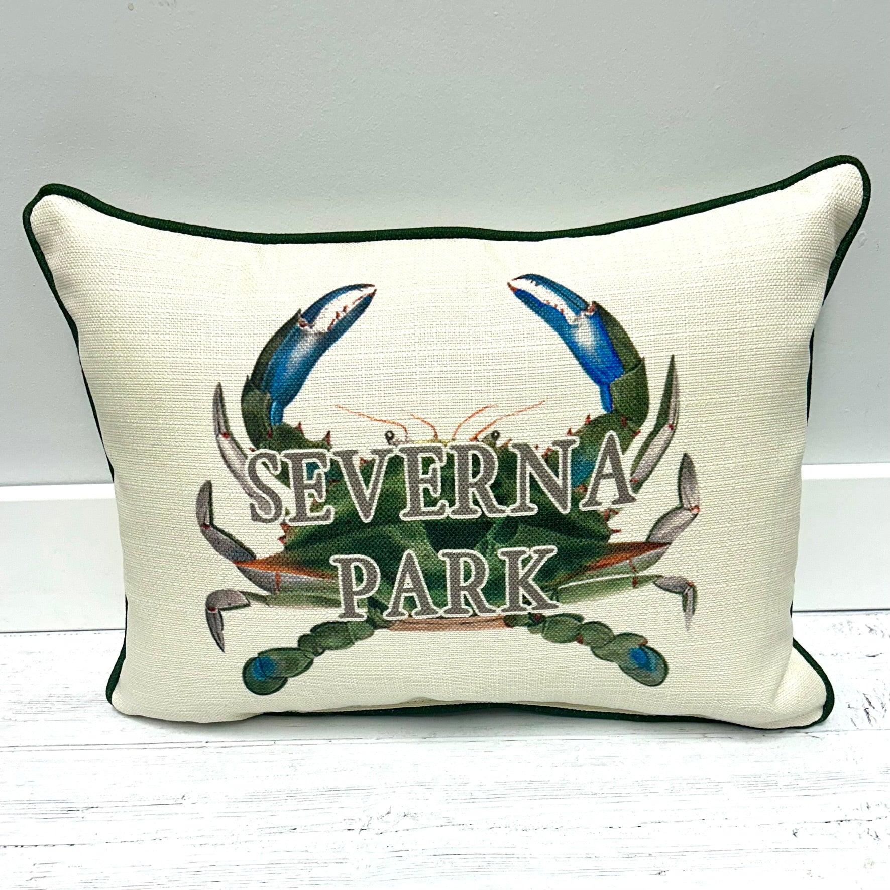 An off-white textured throw pillow with dark green piping, featuring a printed design of a green and blue crab with the words 'SEVERNA PARK' across the front, set against a light gray background.