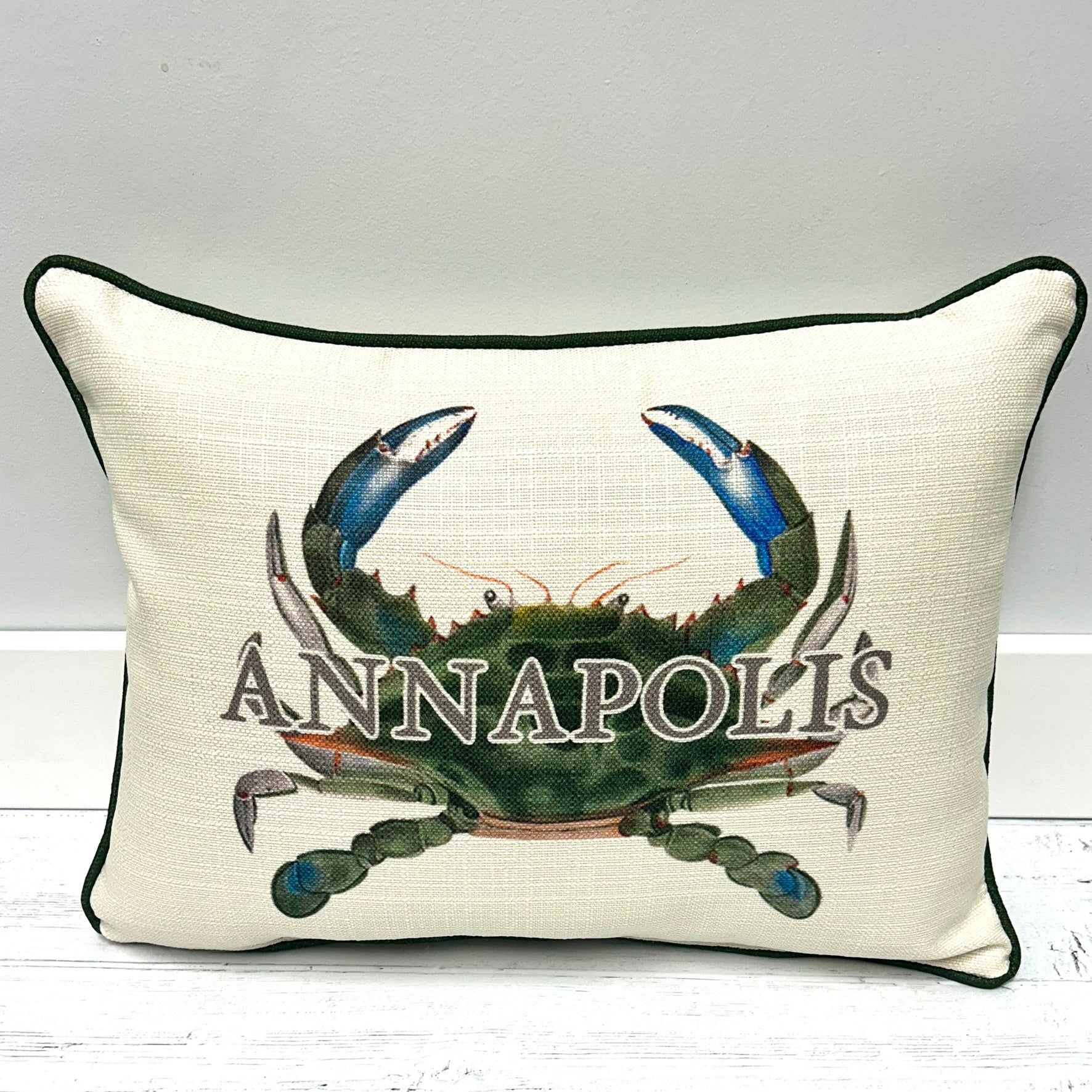 An off-white rectangular throw pillow with dark green piping, featuring a large embroidered blue and green crab and the word 'ANNAPOLIS' across the front.