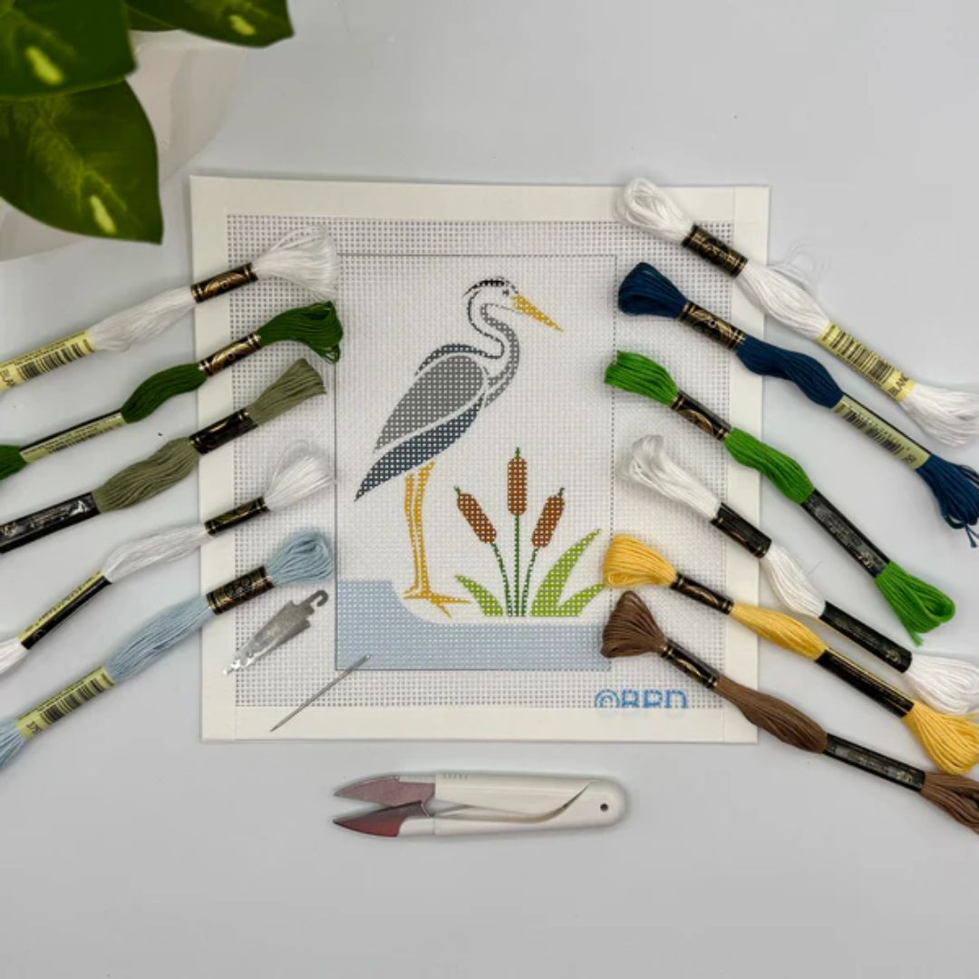 Cross-stitch kit with a heron design, embroidery threads, and tools on a light gray background.