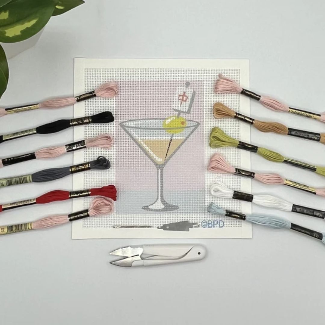 Cross-stitch pattern with a martini glass design, embroidery threads, and a scissor on a light gray background.