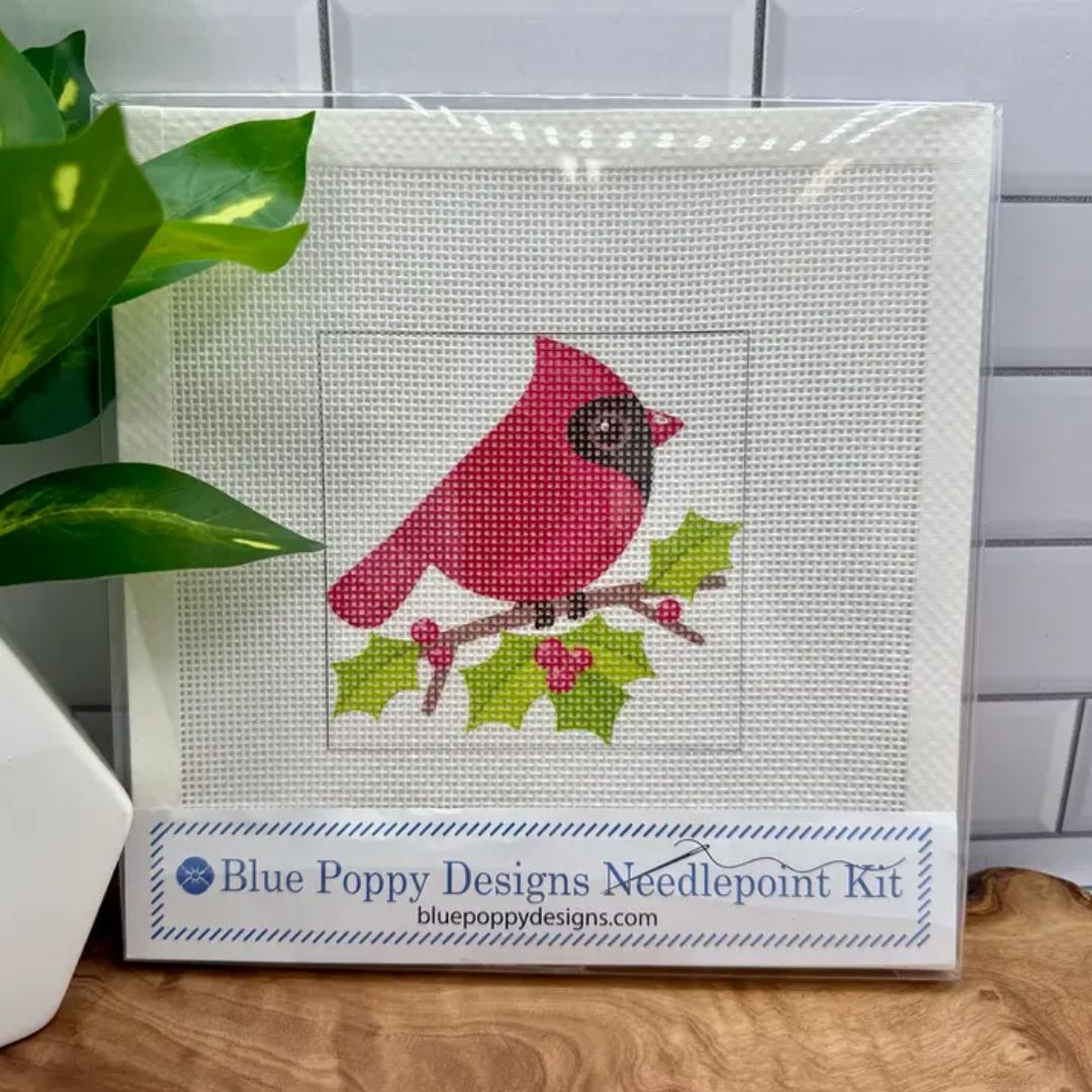 A packaged needlepoint kit showing a cardinal bird with green holly leaves on a white mesh canvas, with a branded label at the bottom reading Blue Poppy Designs Needlepoint Kit. The package is photographed on a wooden surface with a plant and white tile wall in the background.